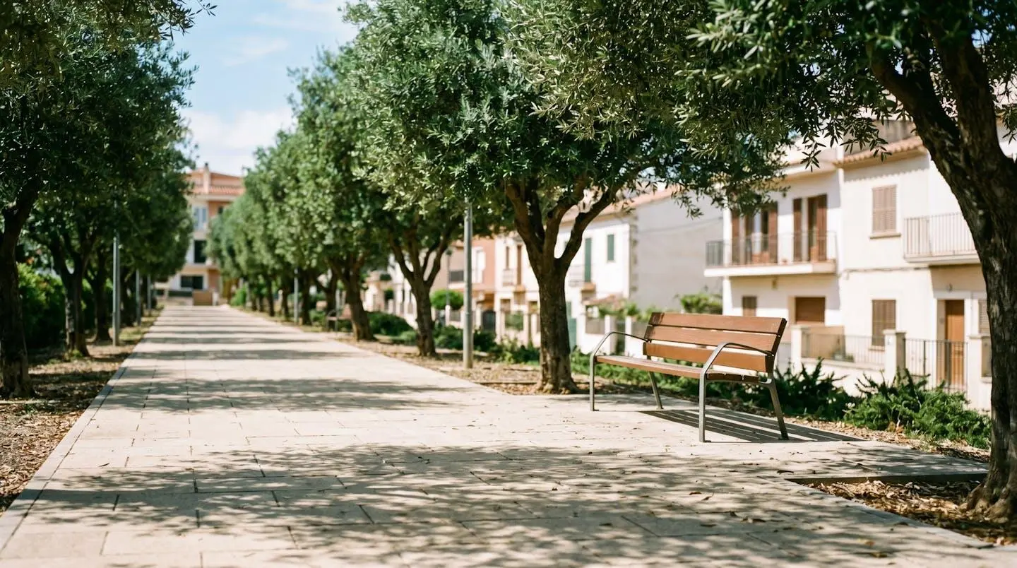 Allée piétonne ombragée bordée d'arbres matures avec pavement clair, banc design au second plan et immeubles résidentiels contemporains en arrière-plan sous lumière filtrée